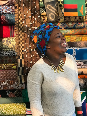 A woman wearing a blue scarf, gold necklace and white sweater. Various colorful fabrics in the background