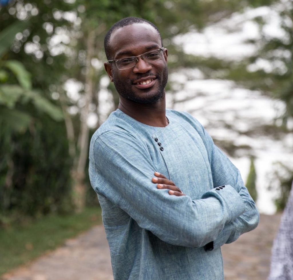 A man wearing glasses and a blue shirt stands with arms crossed, smiling toward the camera.