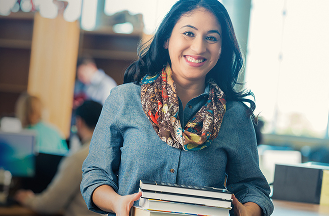 Image of a woman with dark hair holding books dressed in a blue long sleeve button up shirt and a red, yellow print designed scarf.