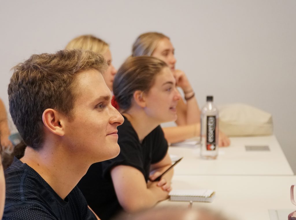 a smiling young male student sits at a table with three female students, listening to a presentation