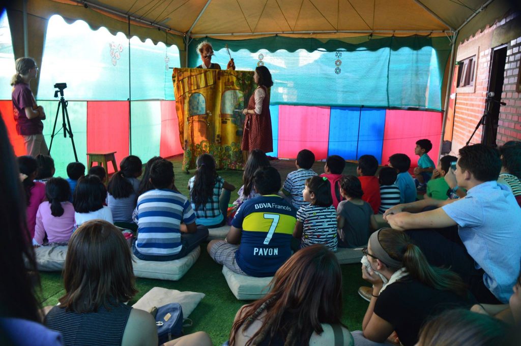 21 children sit on cushions on the floor and watch a puppet show