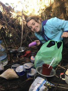 A young woman crouches and smiles near her camp supplies