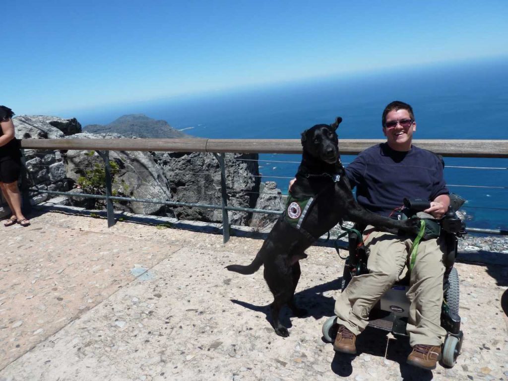 Alex Stone in his wheelchair in front of a railing on a high cliff beside the ocean in South Africa with service dog Fraser beside him