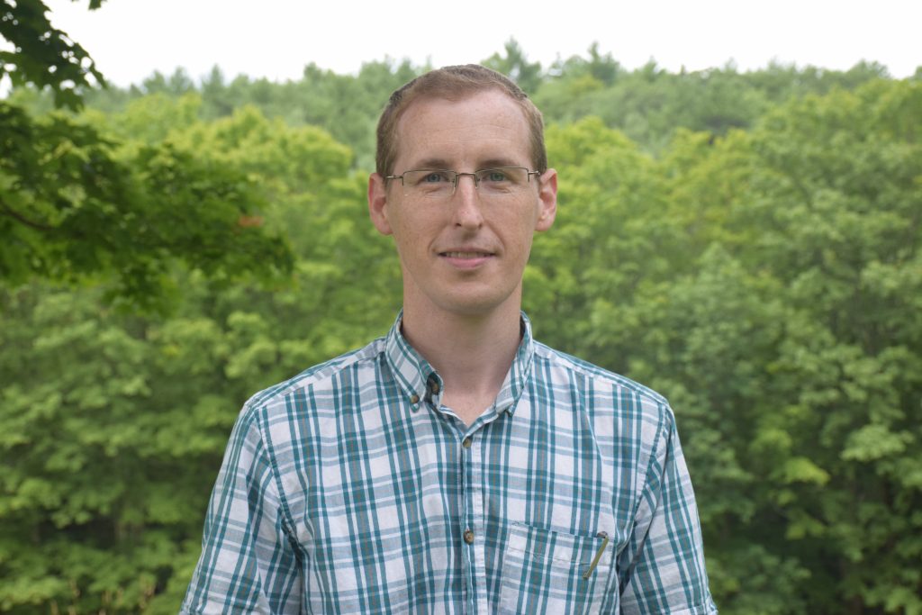 Man wearing glasses and plaid shirt smiling with lots of leafy trees in the background