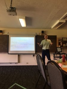 Same man from above presenting next to a projector screen with information projected onto it