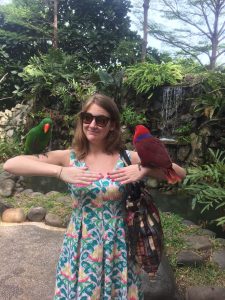 Young woman in sundress and sunglasses poses outside with a tropical bird on each arm