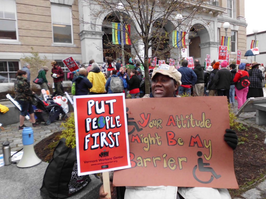 Woman in wheelchair outside with signs that say "Put People First" and "Your Attitude Might Be My Barrier"