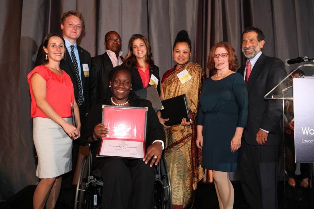 Komabu Pomeyie in wheelchair, holding her award, surrounded by seven people of varying genders, ethnicities, and ages