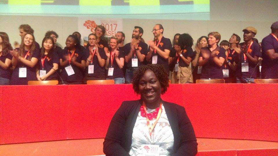 Woman sitting and smiling in the foreground while 25 people applaud behind her