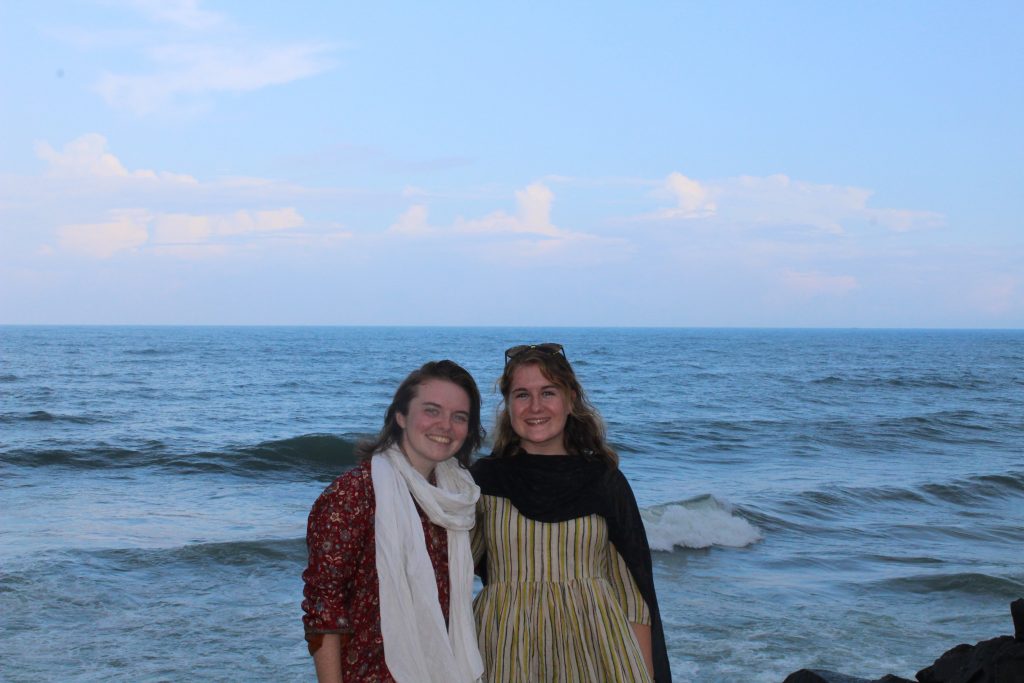 Two young women stand smiling in front of the ocean