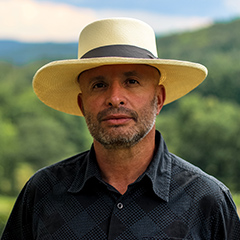 A man with a short beard wearing a broad-brimmed Panama-style hat and a black shirt.