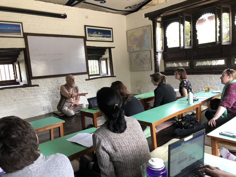 Lecturer seated in a classroom, with students at long green desks