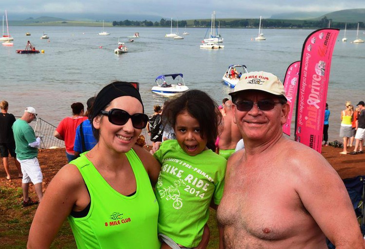 A man, a young woman and a child smile at the camera. Behind them is a body of water with sailboats.