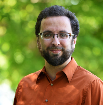 Man with glasses, dark hair and beard in an orange shirt.