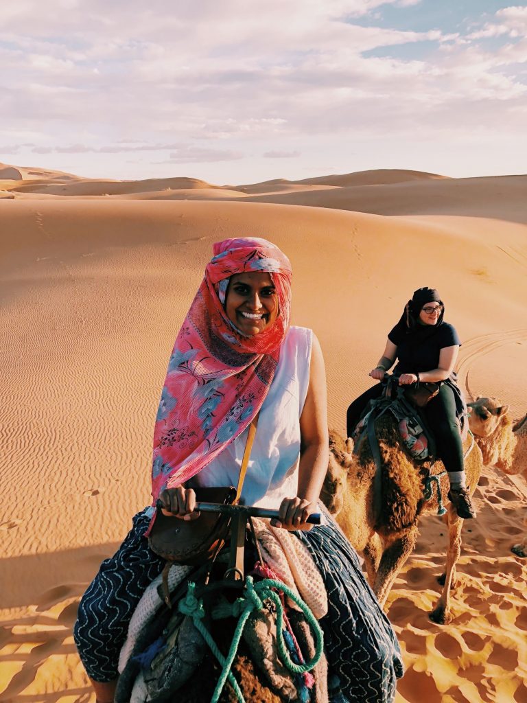 Two students riding camels in the desert