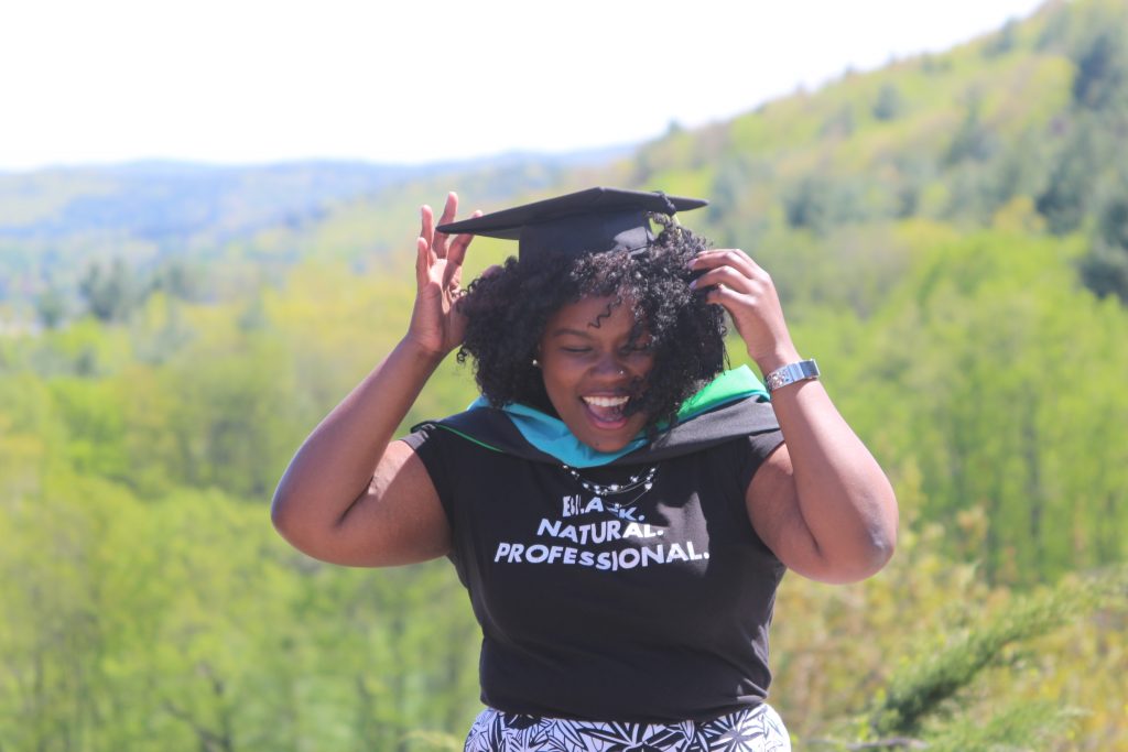 A woman in a graduation cap smiles as she holds the cap. Her T-shirt says "Black. Natural. Professional."