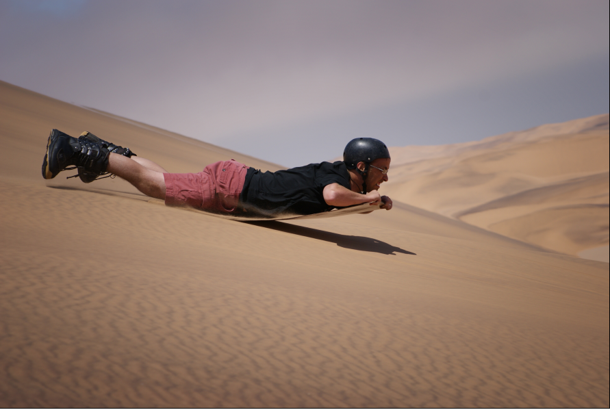 A man in shorts, T-shirt and helmet boogie-boarding down a sand dune.