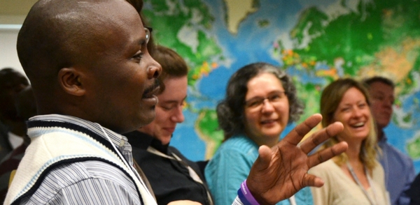 In the foreground, a man in profile gestures while three others in the background smile. On the wall is a large world map.