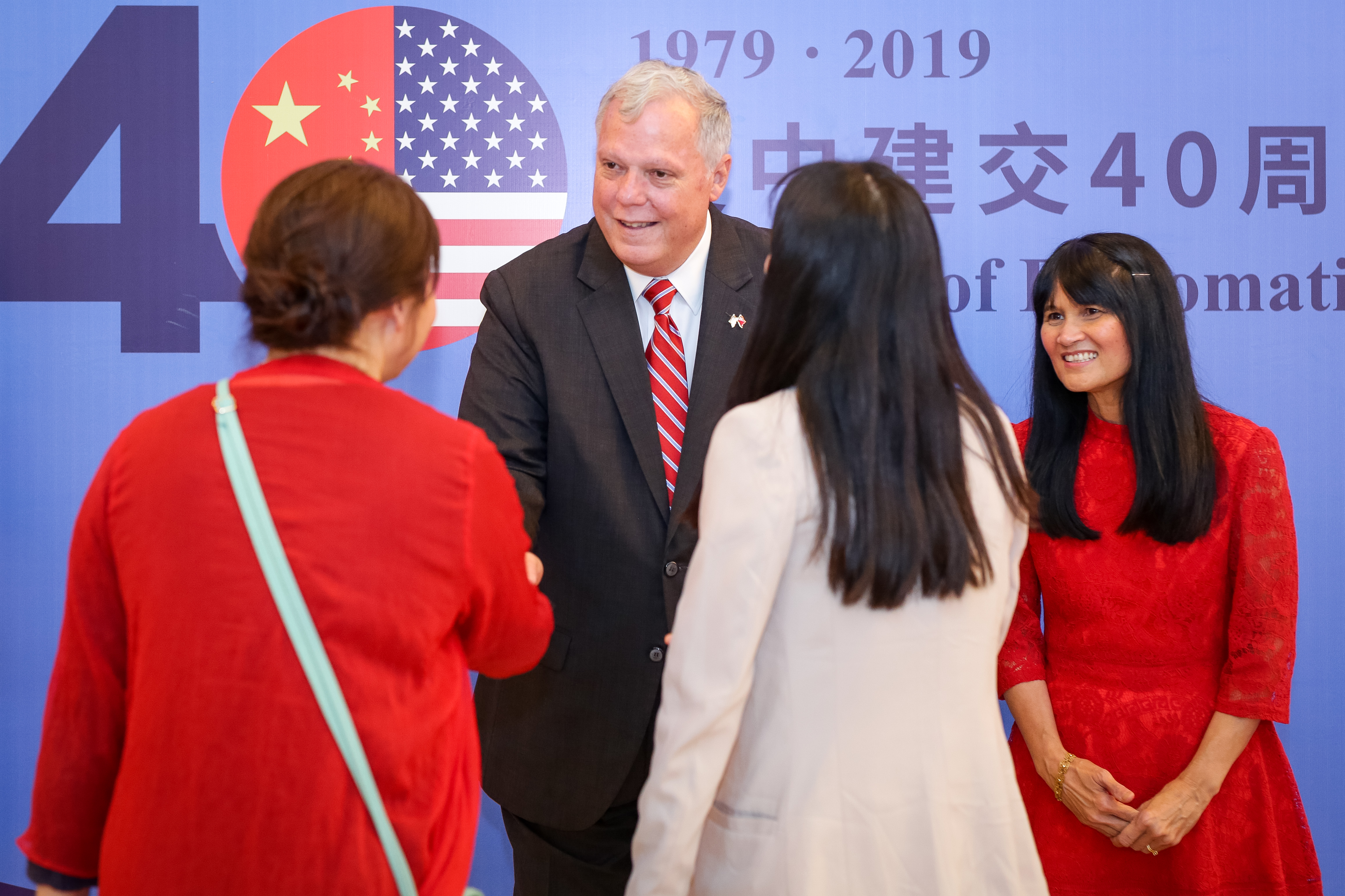 A man in a suit (Jamie Fouss) stands and a woman in a red dress greet two women who are facing away from the camera. In the background a large banner  commemorates the 40th anniversary of U.S.-China diplomatic relations.
