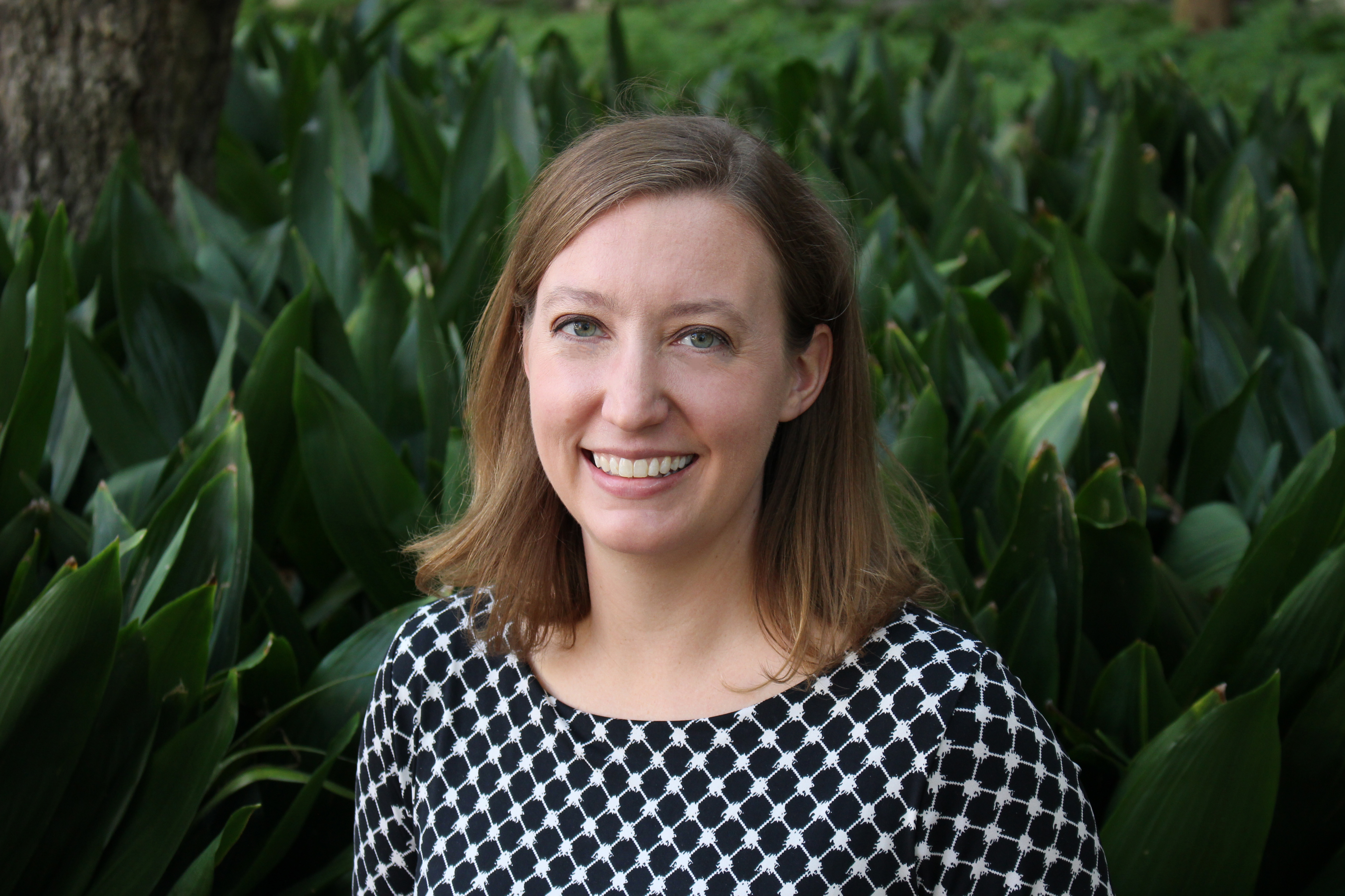 A woman with light brown hair, wearing a black and white blouse, stands against a backdrop of green plants.