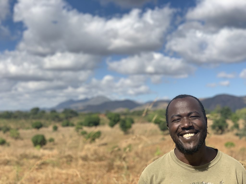 A man is pictured in the lower right corner with a field and mountains behind him.