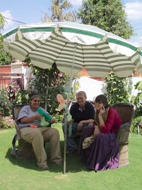 Three young adults sit in wicker chairs under a large sun umbrella two holding balloon flowers.