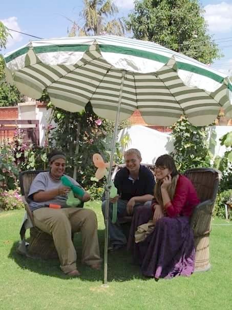 Three young adults sit in wicker chairs under a large sun umbrella two holding balloon flowers. 