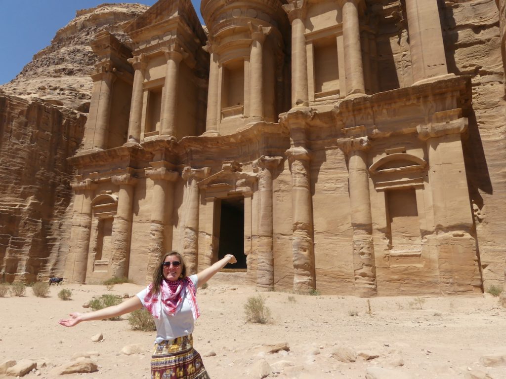 A young woman stands with arms outstreched in front of a building carved into a rock wall in Petra.