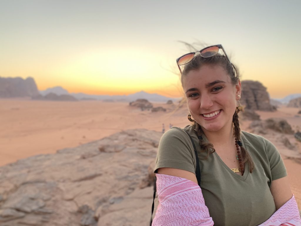 A young woman two braids in her hair smiles at the camera while she stands in Wadi Rum.