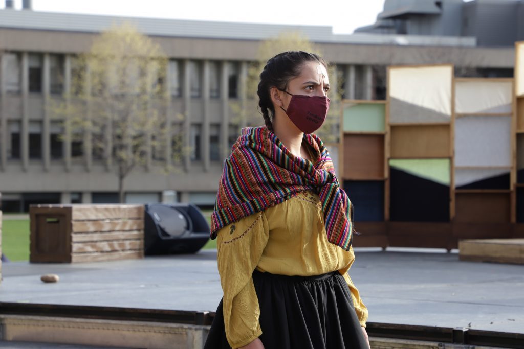 A woman in a yellow blouse, colorful shawl, and a face mask performs on an outdoor stage.