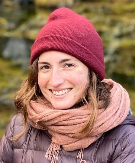 A smiling young woman with brown hair wearing winter clothing: a red cap, mauve scarf and purple jacket