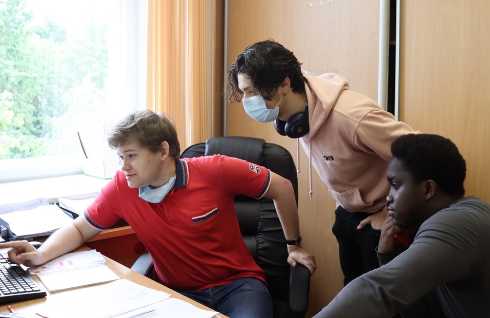 Three men, one wearing a surgical mask, look at a computer screen.