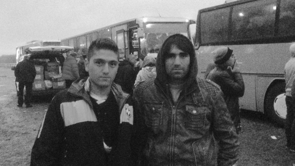 A black and white photo of two Afghan refugees in coats standing near buses in Serbia.