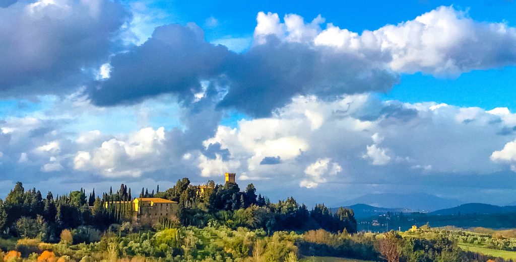 An Italian field and villa atop a hill, against blue skies with white clouds