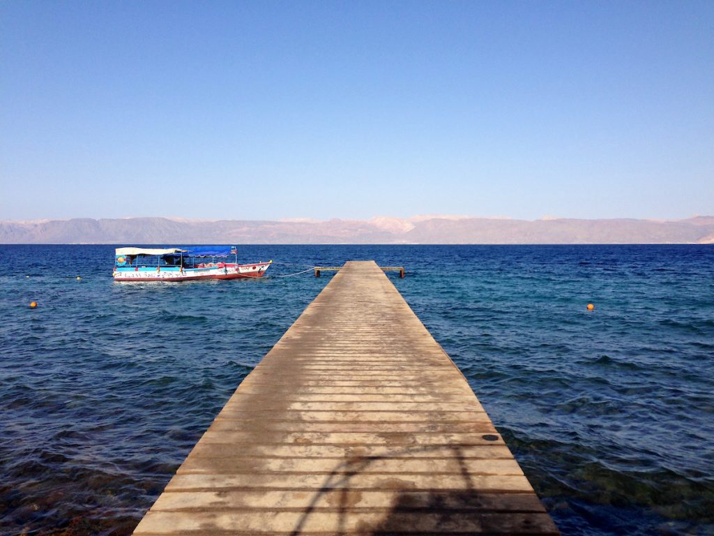 A wooden dock extends into a lake where there is a blue and red platform boat. Desert hills are in the background.