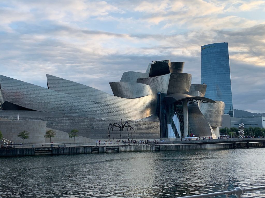 Modern, nonlinear architecture and a statue of a spider on a river bank in Bilbao, Spain.