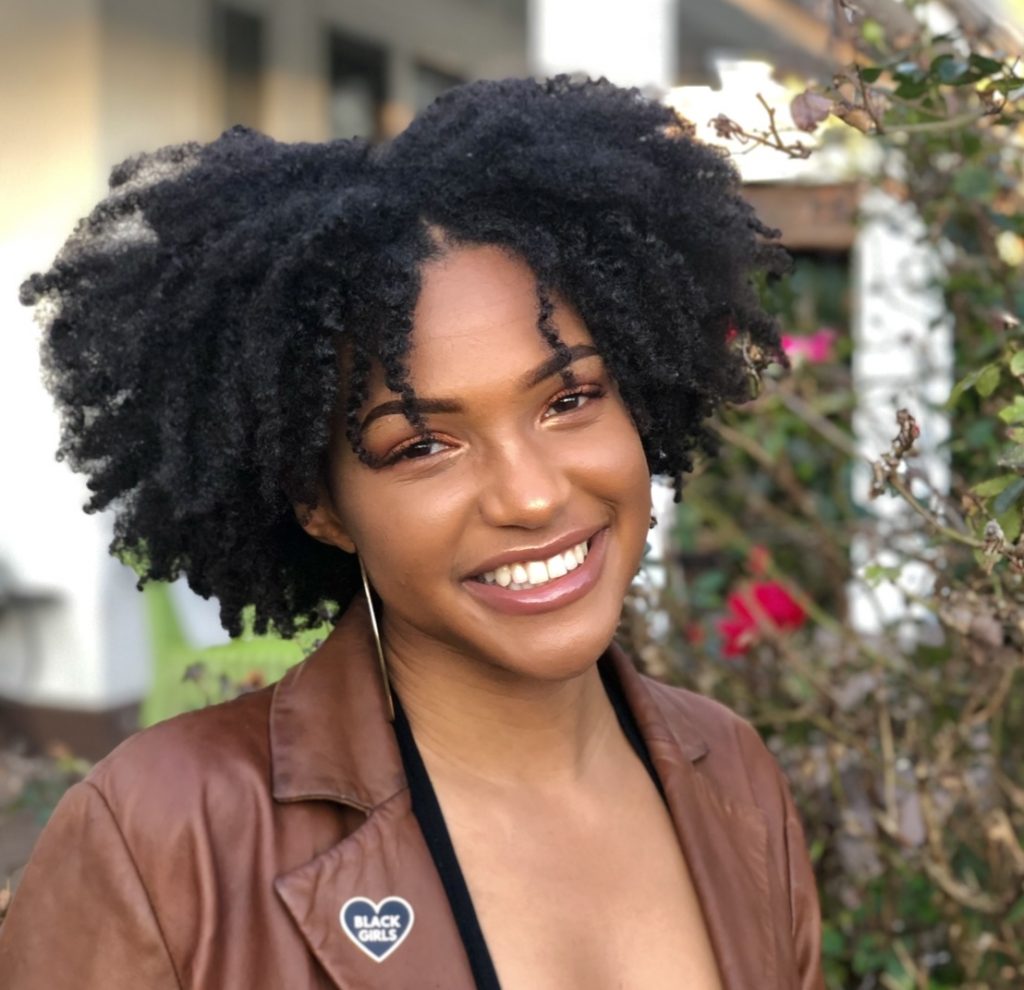 A young woman with short dark hair, silver earrings and a brown jacket smiles at the camera. Her lapel pin is a heart that reads "Black Girls".