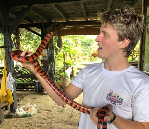 A young man with blond hair and a white t-shirt looks at a large black and orange snake that he's holding.