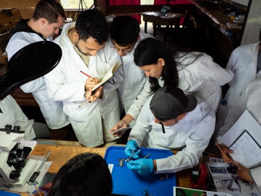 Five students in white lab coats look at specimens in a lab. One is writing in a notebook.