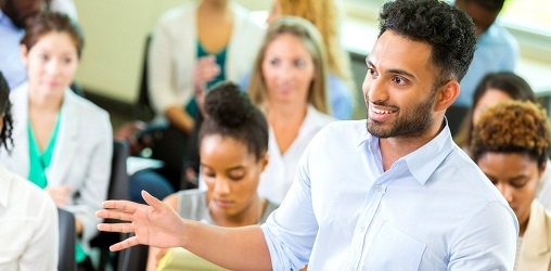 a young man smiles in the foreground with classmates seated behind him.