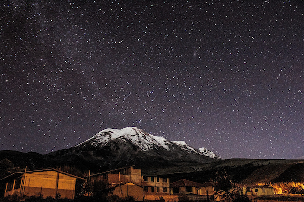 A snow-capped mountain against a starry sky and a rural village in the foreground.