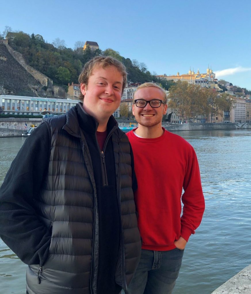 Two young men smile toward the camera. There is a river in the background. The man on the left wears a black vest and black sweater; the one on the right has glasses and a red sweater.