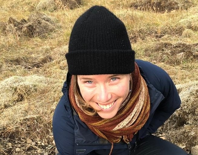 A woman wearing a black knit hat, a brown and gold scarf, and a parka, against a backdrop of dry grass.