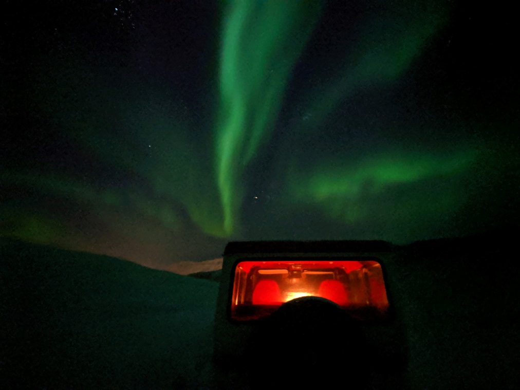 The Northern Lights appear as a green streak in a dark sky. A vehicle in the foreground is lit with and orange interior light.
