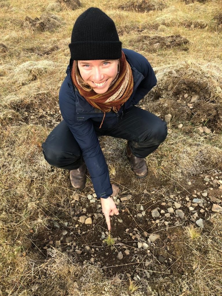 A woman in jacket and black beanie points to a small tree sapling emerging from brown ground.