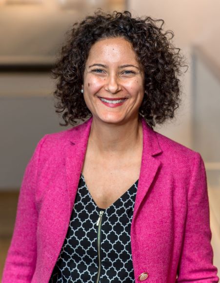A woman with curly dark hard and a pink jacket smiles at the camera