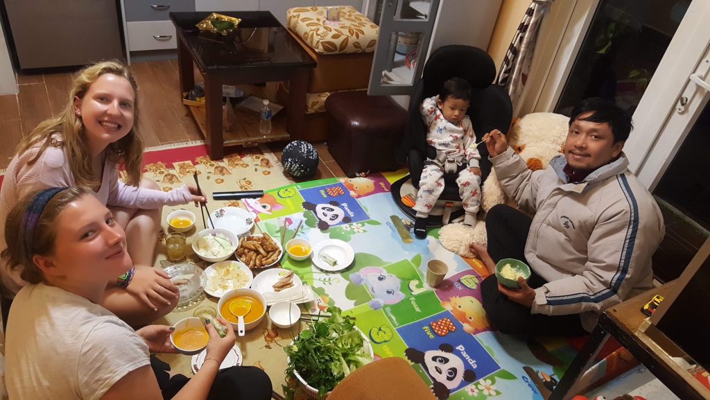 Three adults and a baby gather around a meal set on a cloth on the floor.