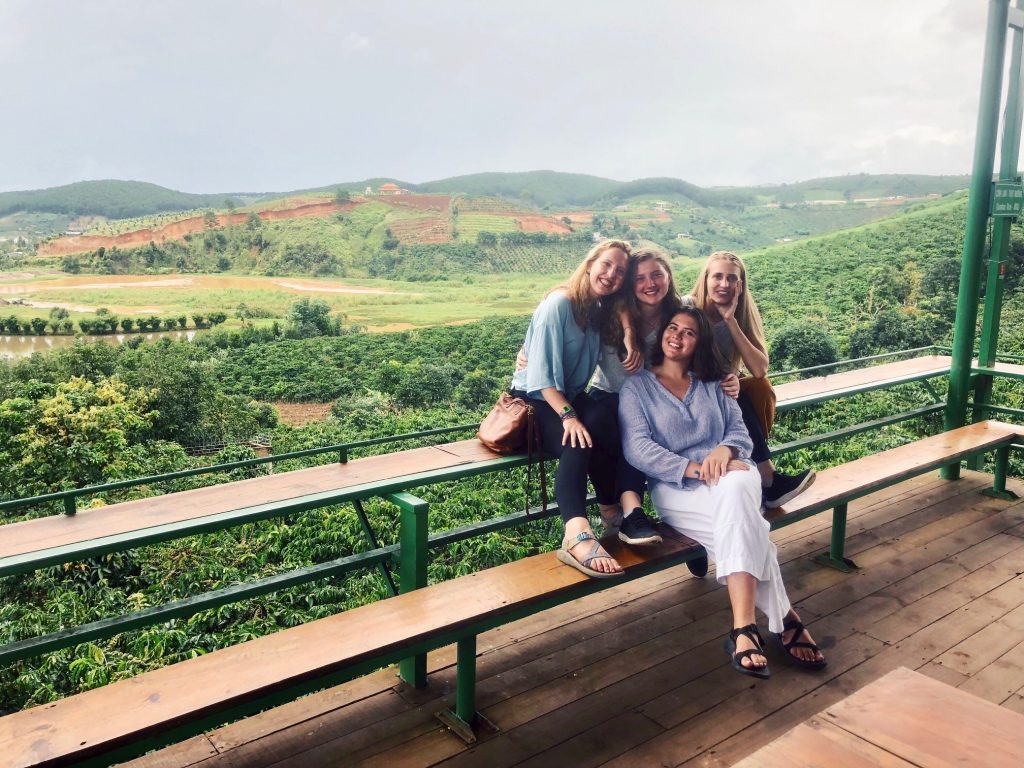 Four young women seated on a bench, smiling toward the camera, with a verdant green countryside in the background