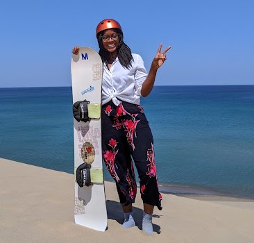 A woman on a beach with a sandboard, giving a V sign.