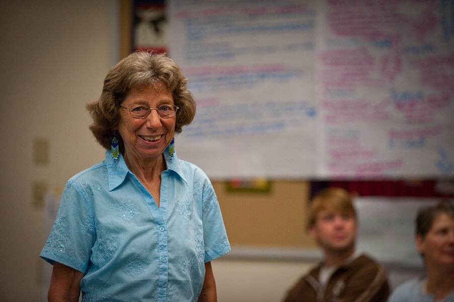 A woman with wire-frame glasses, a light blue shirt and dangly earrings stands against the backdrop of a whiteboard with two students in the background.